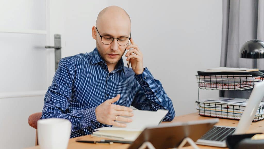 mann sitzt am schreibtisch, während er telefoniert und in einem notizbuch blättert